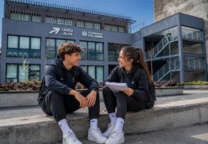 Due studenti seduti all'aperto su un muretto davanti a un moderno edificio scolastico contenti di aver scelto Liceo Alfa.